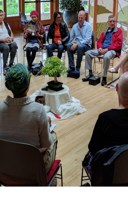 A group of people sit in chairs in a circle. There is a miniature tree in the center on a short table with a table skirt. It is not a pine tree but it does look bonsai'd. 